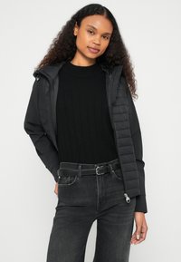 Μαύρο μπουφάν puffer με δομημένο κουκούλι, με καπιτονέ σχέδιο στη μία πλευρά. Φορεμένο πάνω από μαύρο πουλόβερ με ριμπ ύφανση και σκούρο τζιν.