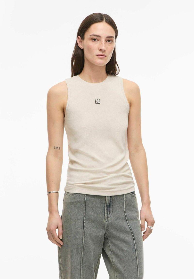 Nő hosszú barna hajjal, bézs színű ujjatlan felsőt visel, melyen kis fekete logó található, valamint laza szürke farmert, fehér háttér előtt áll.