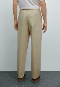 Beige, relaxed-fit trousers with an elastic waistband, two back pockets, and a smooth texture, worn with brown shoes, viewed from the back.