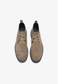 Botas de tobillo de ante marrón con una textura suave, puntera redonda, suela de goma negra y cordones beige. Presenta una pestaña en la parte trasera para facilitar el uso.
