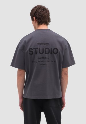 Giovane con capelli castani corti che indossa una maglietta grigio scuro ampia con la scritta nera "WRSTBHVR STUDIO GARMENTS" sul retro.