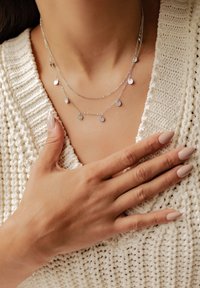 Collier en argent à plusieurs couches avec de petits pendentifs ronds, présenté sur un pull blanc texturé. Les mains reposent sur la poitrine. Texture légère.
