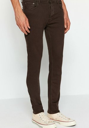 Dunkelbraune Slim-Fit-Jeans, getragen von einer stehenden Person, kombiniert mit elfenbeinfarbenen Leinenschuhen mit Schnürsenkeln.
