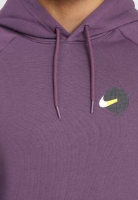 Fialová mikina s texturovaným povrchem, obsahující logo v černé a žluté barvě. Zahrnuje šňůrky se stříbrnými špičkami. Klasický design.