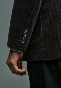 Die Hand eines Mannes neben einem schwarzen Cordjackenärmel mit vier schwarzen Knöpfen, vor einem schlichten grauen Hintergrund.