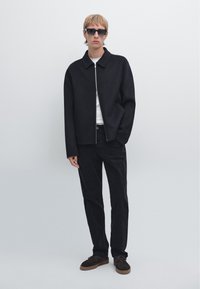 Svart zip-up jacka med krage, bärs över en vit skjorta. Kombinerad med svarta byxor och mörkbruna skor. Modellen står mot en neutral bakgrund.