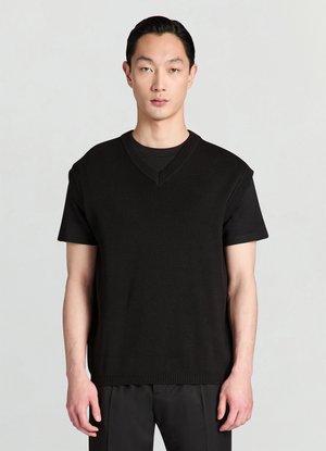 Veste noire en maille avec un col en V et des manches courtes, présentant des accents côtelés au col et à l'ourlet, portée sur une chemise noire.