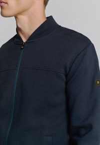 Marine blauwe vest met ritssluiting, gemaakt van gestructureerde stof. Het heeft een opstaande kraag, een turquoise ritssluiting en een hexagonaal logo-accessoire op de mouw.