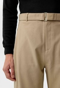 Beige broeken hebben een gladde stof, een rechte snit en een platte voorkant met een metalen sluiting in de taille. Bovenop wordt een zwart shirt met lange mouwen gedragen.
