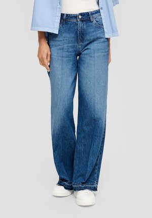 Person wearing loose-fitting blue jeans with raw hems, white sneakers, white top, and light blue open shirt against plain background.