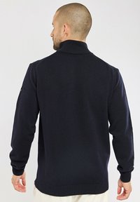 Pull en tricot navy avec un col haut, des poignets et un ourlet côtelés. Présente une texture subtile et un design ajusté, avec des coutures visibles sur les épaules.