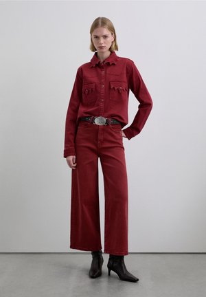 Camisa de mezclilla roja con detalles ondulados y dos bolsillos en el pecho, combinada con jeans rojos de pierna ancha y botines negros con tacón.