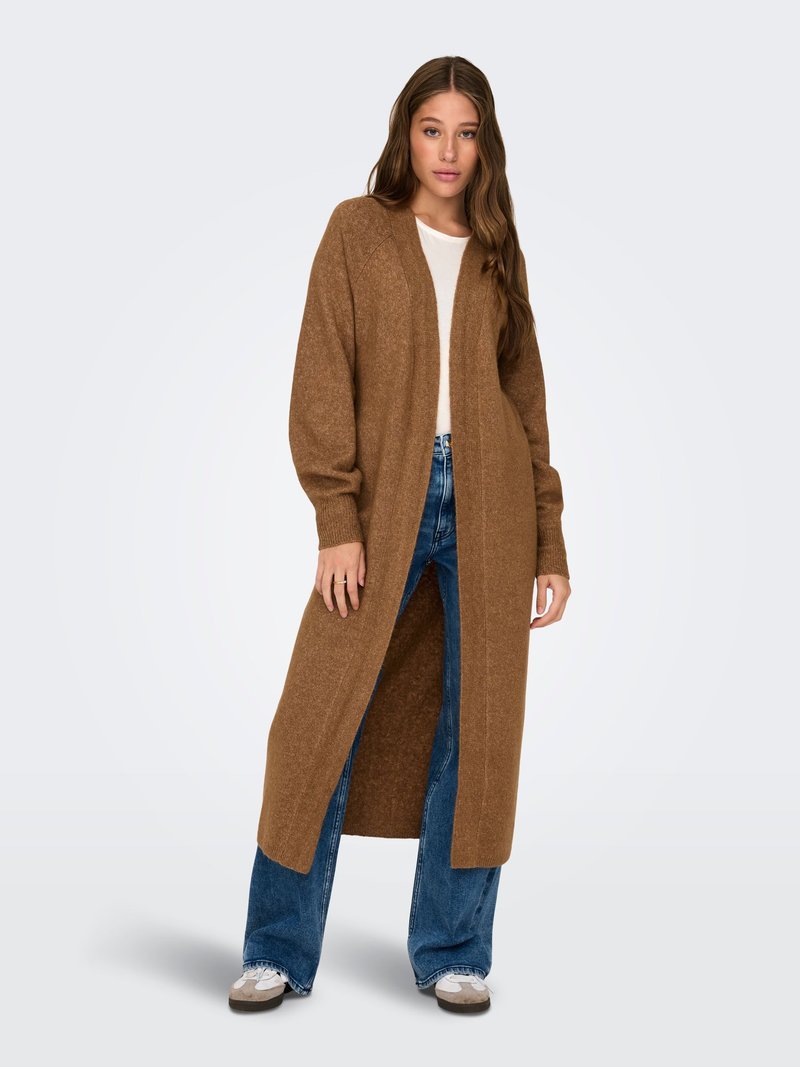 Femme portant un long cardigan marron par-dessus un haut blanc et un jean bleu, debout avec les mains détendues le long du corps devant un fond uni.