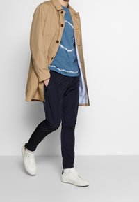 Ljusbeige trenchcoat, blå tie-dye t-shirt, marinblå byxor och vita sneakers med strukturerade sulor. Modellen står i en avslappnad pose.