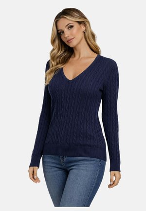 Femme portant un pull en maille torsadée bleu marine à col en V et un jean bleu, debout devant un fond clair uni.