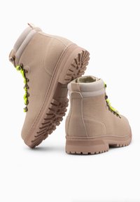 Botas de senderismo beige con suela de goma, puntera reforzada, cuello acolchado y cordones de color verde neón. Presentan una superficie texturizada y un diseño duradero.