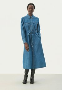 Denim mekko vaaleansinisessä värissä, jossa on kaulus, pitkät hihat, napitettava etuosa, rintataskut ja sidottu vyötärö. Yhdistetty polveen ulottuviin mustiin saappaisiin.