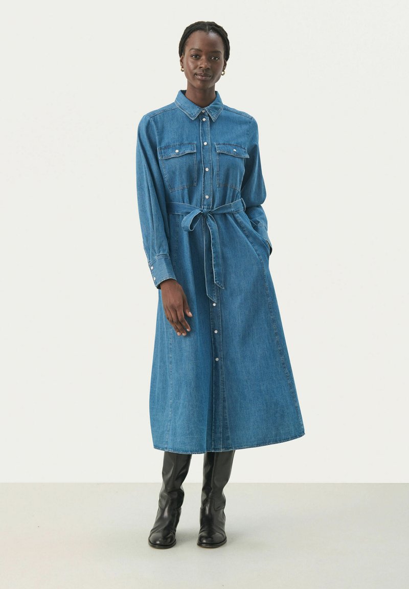 Denim mekko vaaleansinisessä värissä, jossa on kaulus, pitkät hihat, napitettava etuosa, rintataskut ja sidottu vyötärö. Yhdistetty polveen ulottuviin mustiin saappaisiin.