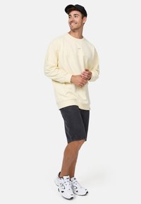 Sudadera amarilla claro con cuello redondo, corte relajado y puños de canalé, combinada con pantalones cortos gris oscuro y zapatillas blancas.