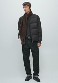 Young man wearing a dark puffer jacket, dark jeans with rolled cuffs, black shoes, and a long brown scarf against a plain light background.