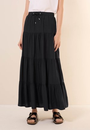 Vrouw draagt een lange zwarte gelaagde rok met een trekkoord in de taille, een wit shirt en zwarte sandalen met gesp, staand tegen een neutrale achtergrond.