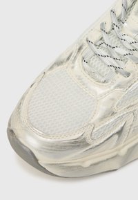 Λευκό αθλητικό παπούτσι με πλέγμα επάνω μέρος και γυαλιστερές λεπτομέρειες, στρογγυλή μύτη και υφασμένη ελαστική σόλα. Σχέδιο κορδονιών σε γκρι με μαύρες βούλες.