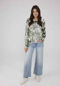 Langärmliges Oberteil mit grün-weißer Musterung und auffälligem Text, kombiniert mit weitgeschnittenen hellblauen Jeans und weißen Sneakers. Sanfte Stofftextur.