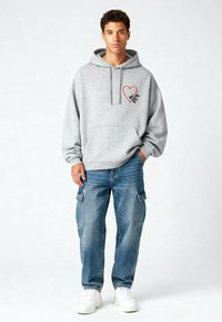 Felpa grigia con tasca frontale e design di cuore rosso. Abbinata a jeans cargo blu e sneakers bianche, in piedi su uno sfondo chiaro.