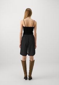 Femme aux longs cheveux blonds portant un haut noir à fines bretelles, un short noir ample et des bottes marron hauteur genoux à talons, de dos.