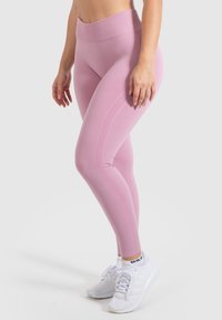 Leggings rosa chiaro con design a vita alta, presentano una cintura a coste e pannelli laterali testurizzati. Indossati con scarpe da ginnastica bianche.