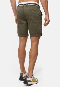 Shorts vert olive avec des revers, deux poches arrière et une ceinture rayée. Portés avec des baskets blanches et grises avec des accents jaunes.