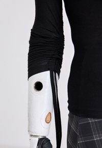 Bílá robotická protetická paže s texturovaným designem, doplněná černým tričkem s dlouhým rukávem, které má shromážděnou látku na rameni.