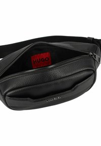 Marsupio in pelle nera con finitura testurizzata, dotato di scomparto principale con zip e etichetta interna rossa. Cintura regolabile inclusa.