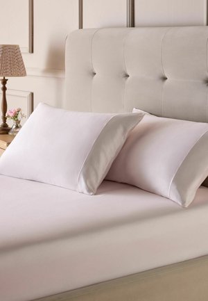 Beiges gepolstertes Bett mit zwei weißen Kissen und passendem Bettlaken, hölzerner Nachttisch mit Lampe und kleiner Vase mit rosa Blumen.