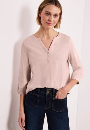 Femme aux cheveux blonds courts portant un chemisier rose clair à boutons avec manches 3/4 et un jean bleu foncé taille haute, main dans la poche.