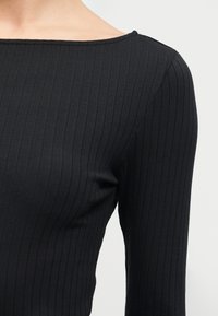 Gros plan d'une personne portant un haut noir côtelé à manches longues avec un décolleté bateau, sur un fond clair uni.