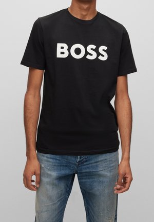 Osoba nosząca czarną koszulkę z krótkim rękawem z napisem "BOSS" dużymi białymi literami oraz niebieskie znoszone jeansy.