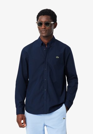 Hombre con camisa azul marino de botones y pantalones azul claro con pequeños logos verdes de cocodrilo, gafas de sol, de pie con una mano en el bolsillo.