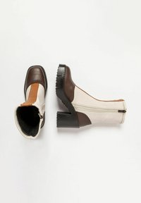 Botas de tobillo de cuero marrón y crema con un tacón grueso negro, que cuentan con un zipper lateral y una textura suave, acentuadas con una franja marrón claro.