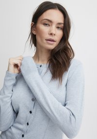 Gaiši zils džemperis, kas izgatavots no mīksta adījuma materiāla, ar pogu aizdari priekšā, ribotām aprocēm un šauru piegriezumu ar redzamām šuvēm.