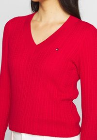 Pull rouge à col en V avec texture côtelée, manches longues et un détail de logo subtil sur la poitrine. Le matériau semble doux et tricoté.