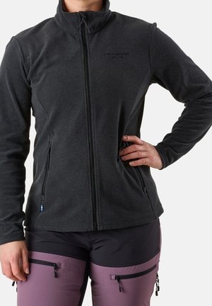 Person trägt eine dunkelgraue Fleecejacke mit Reißverschluss und violette Outdoorhose mit schwarzen Taschen, steht mit einer Hand in der Hüfte.