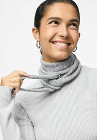 Grå strikket turtleneck med ribbet tekstur, der har et snoet gråt tørklæde omkring halsen, holdt af en hånd. Sølv-hulløreringe er synlige.