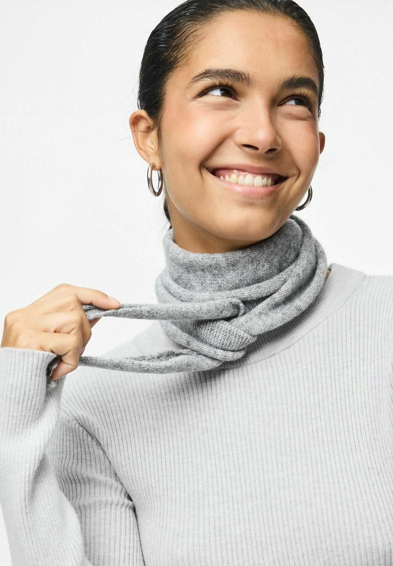 Grå strikket turtleneck med ribbet tekstur, der har et snoet gråt tørklæde omkring halsen, holdt af en hånd. Sølv-hulløreringe er synlige.