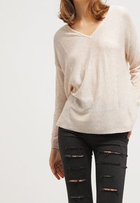 Beżowy sweter o luźnym kroju z dekoltem w serek, z prążkowaną fakturą i zaokrąglonym dołem, zestawiony z czarnymi, podartymi jeansami z licznymi przetarciami.
