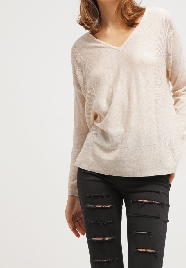 Beżowy sweter o luźnym kroju z dekoltem w serek, z prążkowaną fakturą i zaokrąglonym dołem, zestawiony z czarnymi, podartymi jeansami z licznymi przetarciami.