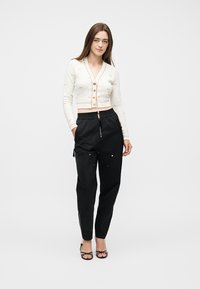 Γυναίκα που στέκεται, φοράει λευκή κοντή ζακέτα με κουμπιά και μαύρο παντελόνι ψηλόμεσο με φερμουάρ και τσέπες, μαύρα σανδάλια με τακούνι.