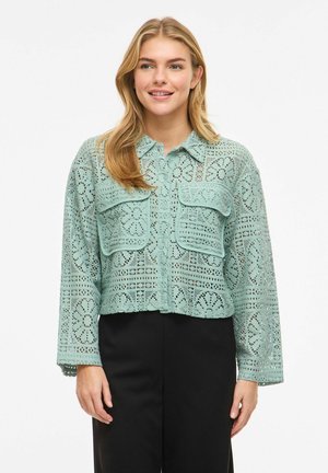 Jeune femme portant une chemise boutonnée en dentelle motif vert menthe avec des poches à rabat à l'avant et un pantalon noir, souriant légèrement, sur fond blanc.