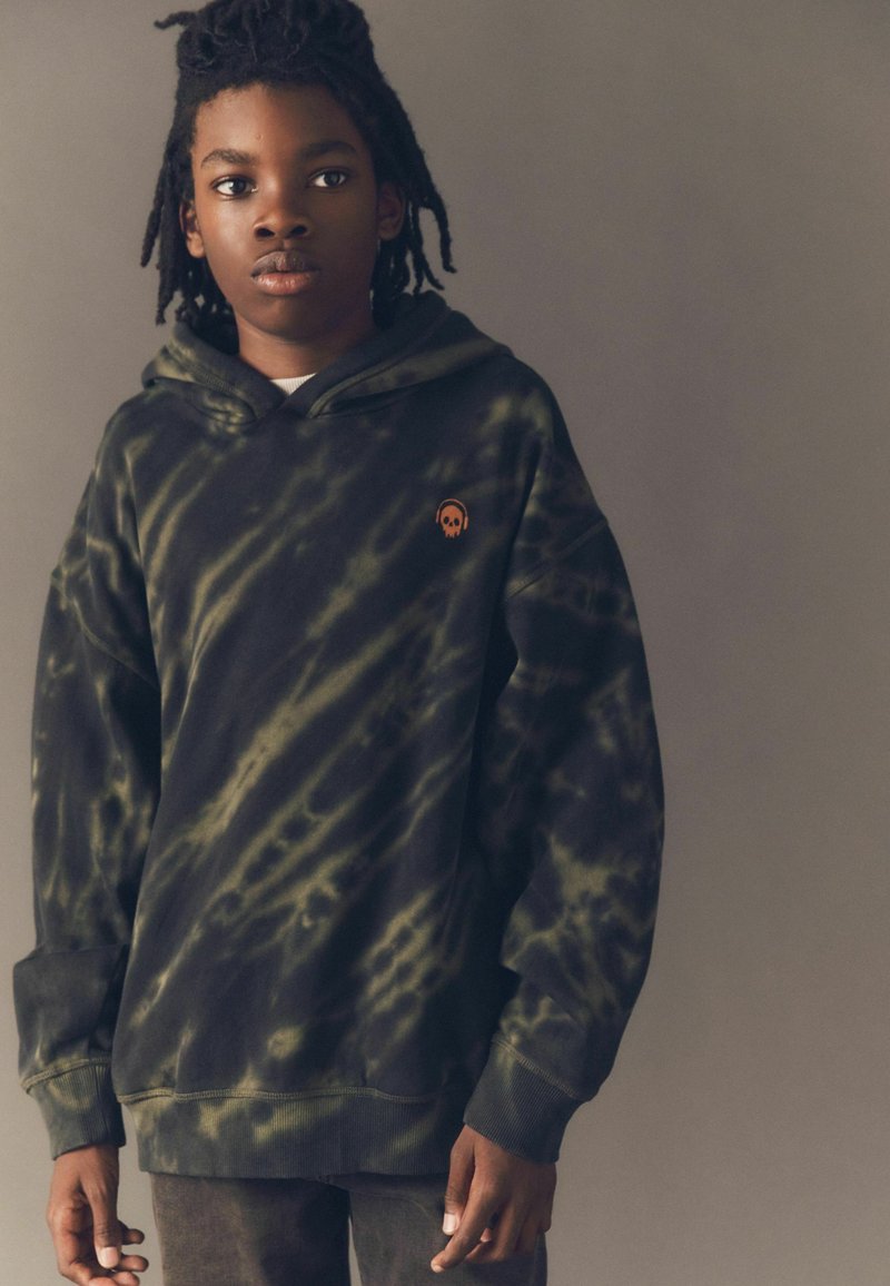 Kapuzensweatshirt in dunklem Grün mit beigem Batikmuster, ausgestattet mit einem orangefarbenen Skull-Logo auf der linken Brust, aus Baumwollmaterial, mit lockerer Passform.