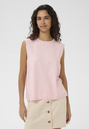 Kobieta o kręconych ciemnych włosach, ubrana w bezrękawnik w jasnoróżowym kolorze oraz beżową spódnicę z guzikami z przodu, na jednolitym tle.